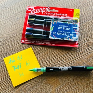 Könnte beinhalten: Eine Packung mit fünf Sharpie Creative Markern in Schwarz mit bunten Akzenten. Die Marker sind wasserbasierte Acrylmarker und haben eine "Paint Effect No Bleed" Funktion. Die Verpackung ist rot und weiß mit dem Sharpie Logo. Ein grüner Sharpie Marker liegt auf einem gelben Haftnotizblock mit dem Wort "hi" mehrmals darauf geschrieben.