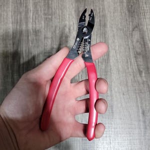 May include: Red handled wire strippers with black cutting blades. The tool is held in a hand and is open. The tool has the text "12-20" and "WARNING SOFT WIRE ONLY" printed on it.