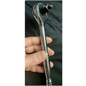 May include: A chrome-plated ratchet wrench with a black square socket attached. The wrench is held in a hand, showing the handle and head of the tool. The tool is designed for tightening or loosening bolts and nuts.