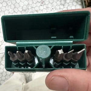 Könnte beinhalten: Eine grüne Kunststoffbox mit einer Reihe verschiedener dunkler Metall-Schraubendreherbits. Die Box ist geöffnet und zeigt die Auswahl an Bits in verschiedenen Formen und Größen, die für verschiedene Schraubentypen geeignet sind. Die Bits sind ordentlich in der Box angeordnet.