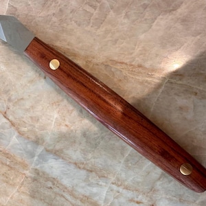 May include: A woodworking knife with a polished, angled steel blade and a long, reddish-brown wooden handle. The handle is secured with two brass rivets. The knife is designed for precision cutting and shaping of wood.
