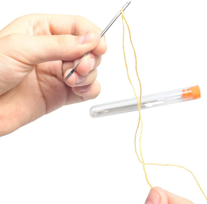 May include: A close-up of a hand threading a needle with yellow thread. The needle is held in a clear plastic tube with an orange cap.