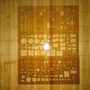 May include: A set of three clear plastic stencils with various shapes and symbols. The stencils are on a wooden surface.
