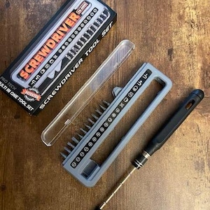 Puede incluir: Un juego de destornilladores multiherramienta con una caja negra y naranja que dice "SCREWDRIVER TOOL SET". El juego incluye un soporte gris con varias puntas y un mango de destornillador negro. El juego está diseñado para uso múltiple.