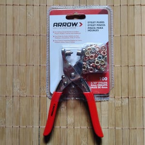 May include: An Arrow brand eyelet pliers set with red handles, packaged with a clear plastic bag of 100 eyelets. The packaging includes text in English and Spanish, and the eyelets are 5/32 inches (4mm).