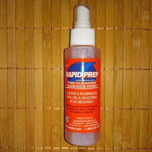 May include: A clear plastic bottle with a white spray nozzle. The bottle is labeled "Rapid Prep Pre Cleaner Surface Prep" and states that it cleans and eliminates wax, oil, and silicones in 60 seconds. The bottle is on a brown bamboo surface.