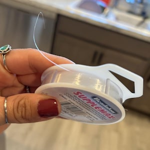 May include: A spool of clear, transparent beading thread with a white label that reads "Beadalon SUPPLEMAX CORDON TRANSPARENT MONOFILAMENTO DE ALUSION CODON 0.15 mm (50 m)"