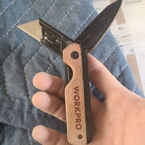 Puede incluir: Un cúter WORKPRO con mango de madera y hoja negra. El cuchillo tiene una hoja retráctil y un mecanismo de empuje. El logotipo WORKPRO está grabado en el mango.
