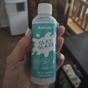Puede incluir: Una botella de plástico transparente de barniz brillante Aukney Clay Glaze, 100 ml, sostenida en una mano. La etiqueta es de color turquesa con texto e iconos blancos que indican secado rápido, mayor brillo y resistencia al agua. La botella tiene una tapa blanca.