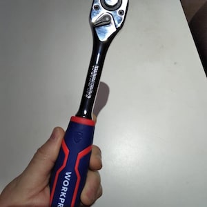 Puede incluir: Una llave de carraca WORKPRO cromada y negra con un mango azul y rojo. La llave se sostiene en una mano y está orientada hacia la esquina superior derecha. El logotipo de WORKPRO está impreso en el mango.