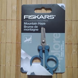 May include: A blue folding pocket scissors with a silver blade. The packaging features the Fiskars logo and the text "Mountain Haze" and "Brume de montagne". The text "Explore" is partially visible on the packaging.
