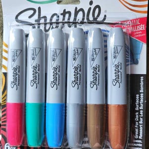 May include: A pack of six Sharpie metallic permanent markers. The markers have silver barrels and coloured tips, including pink, green, blue, grey, gold, and bronze. The packaging includes the Sharpie logo and text indicating the markers are for dark surfaces.
