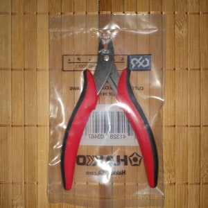 Op de afbeelding: Draadknippers met rode en zwarte handvatten in een doorzichtige plastic zak. De zak heeft een barcode en de tekst "HAKKO" en "HAKKO.com" erop gedrukt.