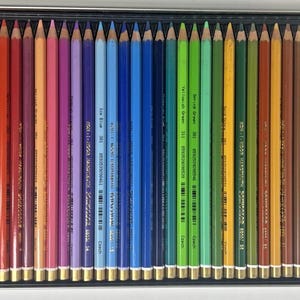 Op de afbeelding: Een set kleurpotloden in een metalen doos. De potloden zijn in een rij gerangschikt en tonen een spectrum van kleuren van geel tot paars. Elk potlood heeft een goudkleurige huls en een label met de kleurnaam en het nummer.
