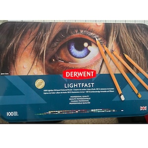 May include: A rectangular tin of Derwent Lightfast coloured pencils. The tin features a detailed eye illustration and the brand name. Four pencils are placed on top. The tin reads "100" and "Professional Quality."