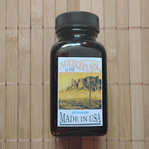 以下が含まれることがあります： 南西部の夕焼けの色をした、90mlのNoodler's Inkのボトルです。ボトルは黒で、ラベルにはサボテンと山のある砂漠の風景が描かれています。ラベルには「pH Neutrality Made in USA」の文字も書かれています。