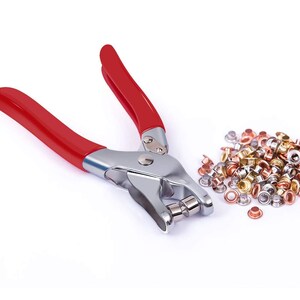 May include: A red handled metal tool for setting eyelets and rivets. A pile of assorted metal eyelets and rivets in silver, gold, and copper tones.