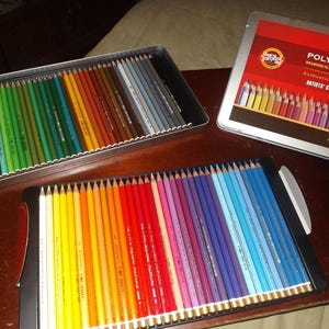 Op de afbeelding: Drie open metalen koffers met kleurpotloden. Eén koffer is open en bevat een regenboog aan kleuren. De andere twee koffers zijn ook open en bevatten een verscheidenheid aan kleuren. De koffers staan op een bruin oppervlak.