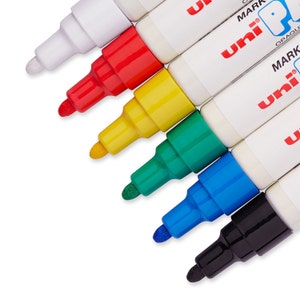 Könnte beinhalten: Ein Satz von acht Permanentmarkern mit deckender Tinte. Die Marker sind weiß, rot, gelb, grün, blau und schwarz. Die Marker sind fächerförmig angeordnet, wobei die Spitzen zum Betrachter zeigen. Die Marker sind mit "uni MARK OPAQUE" beschriftet.