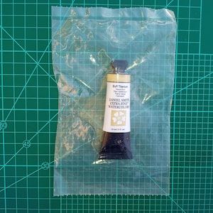 May include: A tube of Daniel Smith Extra Fine Watercolors, labelled "Buff Titanium," is sealed in a clear plastic bag. The tube is cream and dark brown, with text indicating 15 ml (0.5 fl. oz). The background is a green cutting mat.
