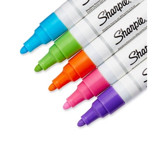 Könnte beinhalten: Nahaufnahme von fünf Sharpie-Markern in leuchtenden Farben: Blau, Grün, Orange, Pink und Lila. Die weißen Stifte tragen das Sharpie-Logo und Text. Die Spitzen der Marker sind sichtbar und zeigen ihre kräftigen Farben.