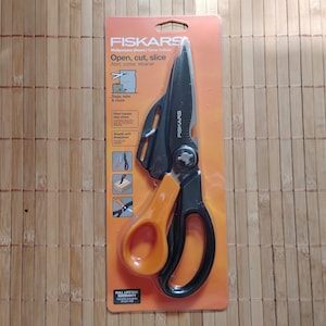 Könnte beinhalten: Schwarze und orangefarbene Fiskars-Mehrzweck-Schere mit einer gezahnten Klinge und einem integrierten Flaschenöffner. Die Verpackung zeigt den Text "Open, cut, slice" und "Full Lifetime Warranty".