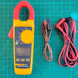 Op de afbeelding: Een gele en rode Fluke 324 True RMS stroomtang met zwarte en rode testkabels. De meter heeft een digitaal display en diverse functieknoppen. De tekst "FLUKE 324 TRUE RMS CLAMP METER" staat op de meter.