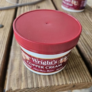 Puede incluir: Un tarro de Wright's Copper Cream con tapa roja y etiqueta blanca. La etiqueta presenta el nombre del producto y las palabras "Trusted Since 1875". El tarro está sobre una superficie de madera. El tarro contiene 227g de producto.