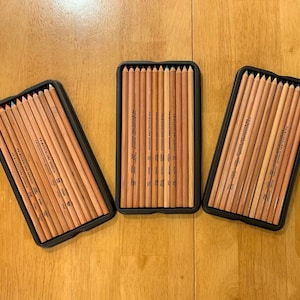 Może przedstawiać: Trzy czarne prostokątne etui wypełnione ołówkami Prismacolor. Ołówki ułożone są w równych rzędach, ukazując naturalny kolor drewna. Etui umieszczone są na drewnianym tle.