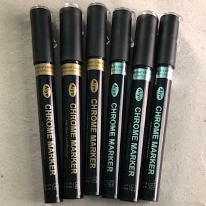 Puede incluir: Seis rotuladores de cromo negro con puntas doradas y plateadas. Los rotuladores tienen el texto "Aiye Chrome Marker" y "High Gloss Liquid Chromium" impreso en ellos.