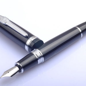 May include: A black fountain pen with silver accents and a fine nib. The pen is lying on a white surface.