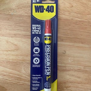 Puede incluir: Un bolígrafo de precisión WD-40 en una tarjeta azul y amarilla. El bolígrafo tiene una tapa roja y detalles en blanco y amarillo. La tarjeta dice "NEW!" y "ORIGINAL WD-40 FORMULA ON-THE-GO". Está diseñado para espacios reducidos y proyectos pequeños.