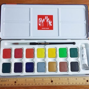 Puede incluir: Juego de pintura de acuarela metálica abierta con 12 colores vibrantes, un pincel y un tubo de pintura blanca. La tapa presenta el logotipo "Caran d'Ache" y el texto "Swiss Made". Una regla de madera está debajo.