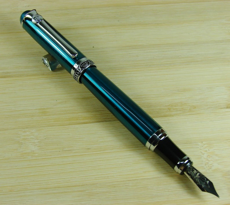 May include: A teal blue fountain pen with silver accents and a fine nib. The pen has a decorative band with a floral design.