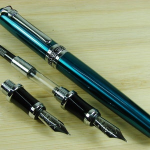 May include: A teal blue fountain pen with silver accents and a clear converter. The pen is lying on a wooden surface with two black and silver fountain pen nibs.