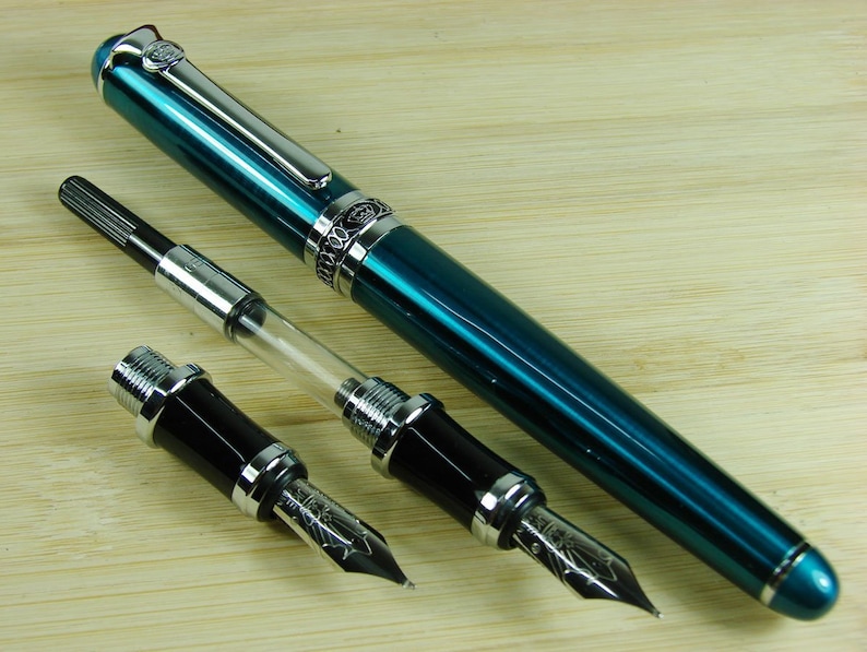 May include: A teal blue fountain pen with silver accents and a clear converter. The pen is lying on a wooden surface with two black and silver fountain pen nibs.