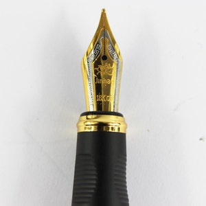 May include: Close-up of a gold-plated fountain pen nib with a black resin body. The nib is engraved with the words "Jinhao 18K GP".