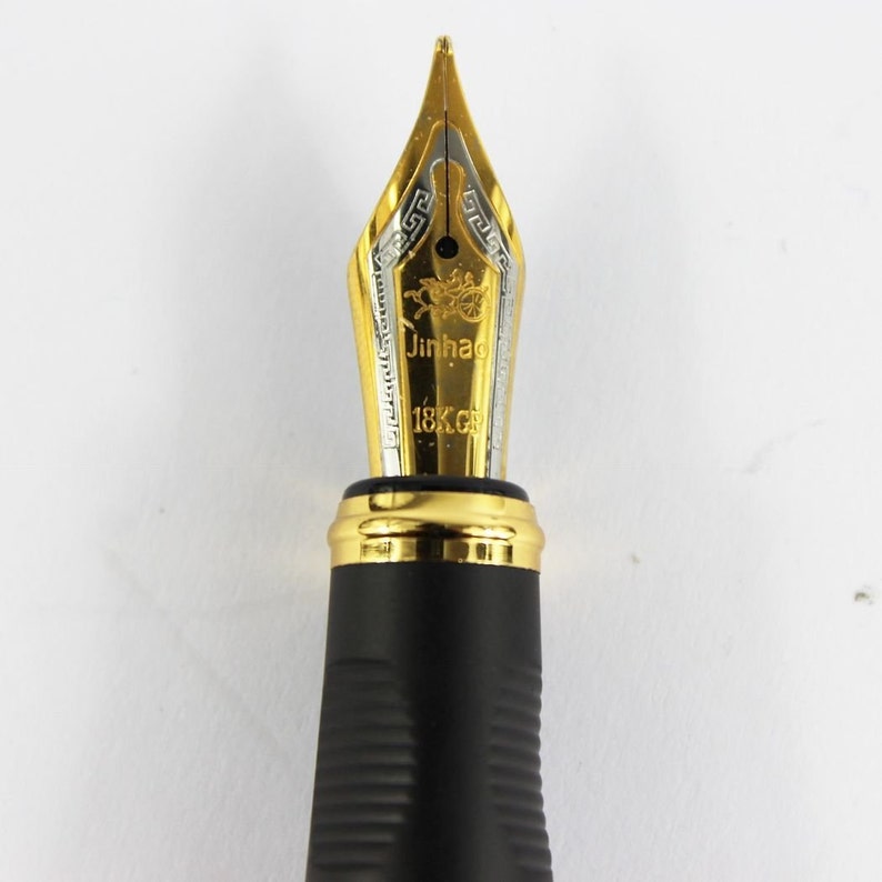 May include: Close-up of a gold-plated fountain pen nib with a black resin body. The nib is engraved with the words "Jinhao 18K GP".