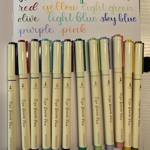 Puede incluir: Colección de rotuladores Sign Brush Pen en varios colores, incluyendo negro, azul, verde, rojo, amarillo y morado. Los rotuladores tienen un cuerpo color crema con el texto "Sign Brush Pen" y "WRITECH". La imagen también muestra nombres de colores escritos en cursiva.