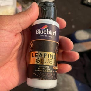 May include: A bottle of Bluebird Leafing Glue, a non-toxic water-based gilding adhesive. The bottle is white with a black cap and a label featuring the Bluebird logo and product information. The label has gold and dark blue accents.