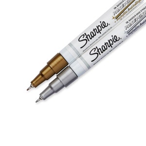 Könnte beinhalten: Zwei Sharpie-Farbstifte, einer goldfarben und einer silberfarben, mit dem Markennamen und der Produktbeschreibung auf dem Schaft. Die ölbasierten Farbstifte sind metallisch.