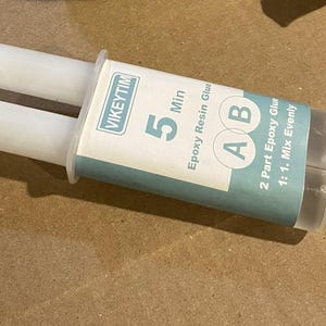 May include: A white and clear epoxy resin glue applicator with the text "VIKEYTIM 5 Min Epoxy Resin Glue". The applicator has two chambers and a mixing nozzle. The label also reads "2 Part Epoxy Glue 1:1. Mix Evenly".