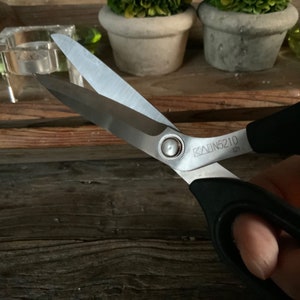 Może przedstawiać: Para nożyczek z czarnym uchwytem i srebrnymi ostrzami. Nożyczki mają czarny uchwyt ze srebrnym pierścieniem i tekst "KAIDN5210" na ostrzu.