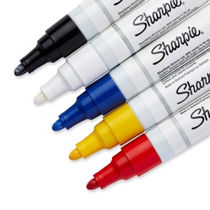 Könnte beinhalten: Ein Set mit sechs Sharpie Permanentmarkern in Schwarz, Weiß, Blau, Gelb, Rot und Silber. Die Marker sind fächerförmig angeordnet, wobei die Spitzen zum Betrachter zeigen. Die Marker sind alle in ihrer Originalverpackung und haben das Sharpie-Logo an der Seite.