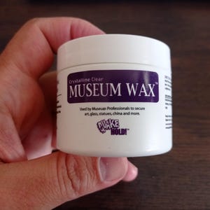 Op de afbeelding: Een witte pot met een paars etiket waarop "Crystalline Clear MUSEUM WAX" staat. Het etiket vermeldt ook "Used by Museum Professionals to secure art, glass, statues, china and more." en "QUAKE HOLD!"