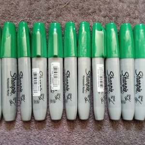 May include: A close-up of twelve green Sharpie permanent markers with chisel tips. The markers are arranged in a row on a gray surface.