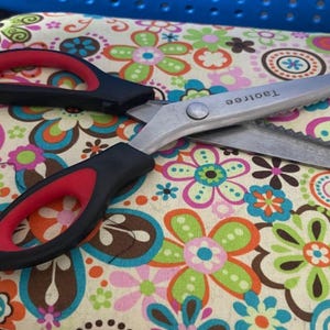 May include: A pair of red and black fabric scissors with a serrated blade. The scissors are on a floral fabric background. The brand name "TaoTree" is printed on the scissors.