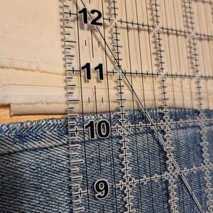 May include: A clear ruler with inch markings, measuring up to 13 inches, is positioned over denim fabric and cream-coloured fabric. The ruler is angled, intersecting the fabrics. The denim is blue, and the cream fabric is layered.