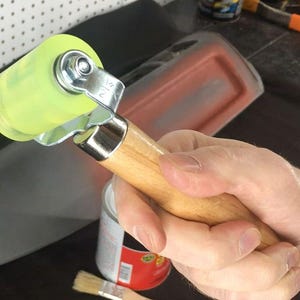 May include: A hand tool with a light green roller and a wooden handle. The roller is attached to a metal frame. The handle is made of light-colored wood. The tool is designed for applying pressure.