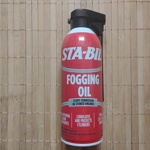 Peut inclure: Une bombe rouge et blanche d'huile de brouillard Sta-Bil. La bombe a un bouchon noir et une étiquette blanche avec le texte "Sta-Bil Fogging Oil" en grandes lettres rouges. L'étiquette indique également "Stops corrosion in stored engines" et "Lubricates and protects cylinders".